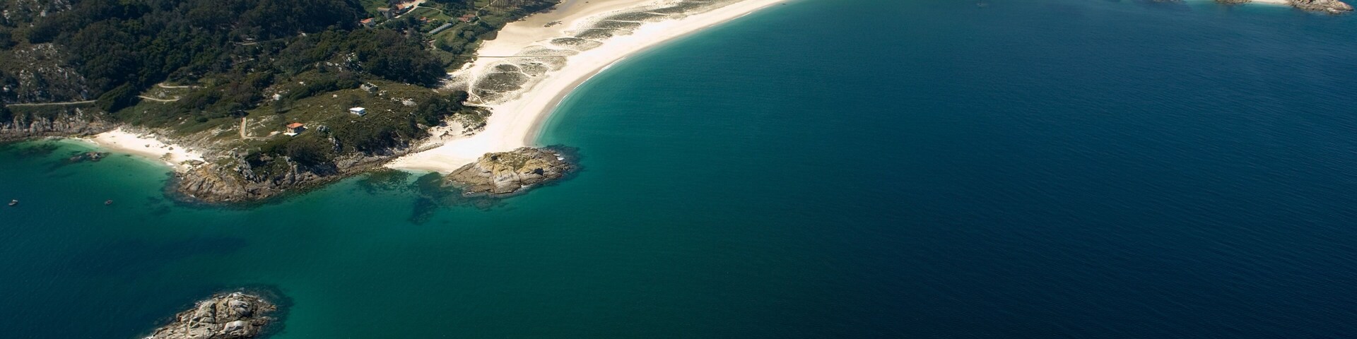 Vista aérea de un paisaje costero con una playa de arena, aguas turquesas cristalinas y un promontorio rocoso destacado.