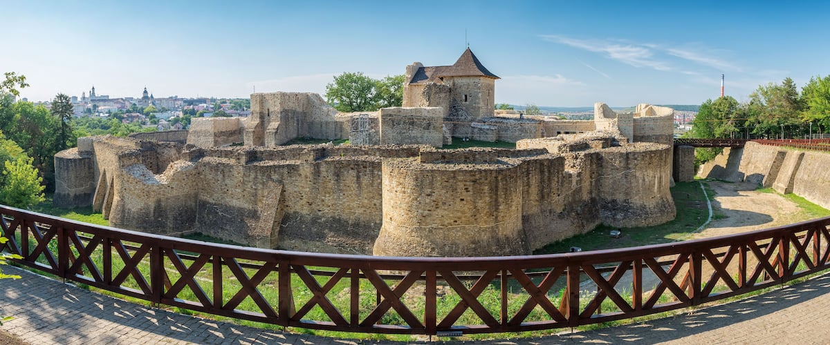 A historic stone fortress with a central tower, surrounded by a moat and a wooden fence.