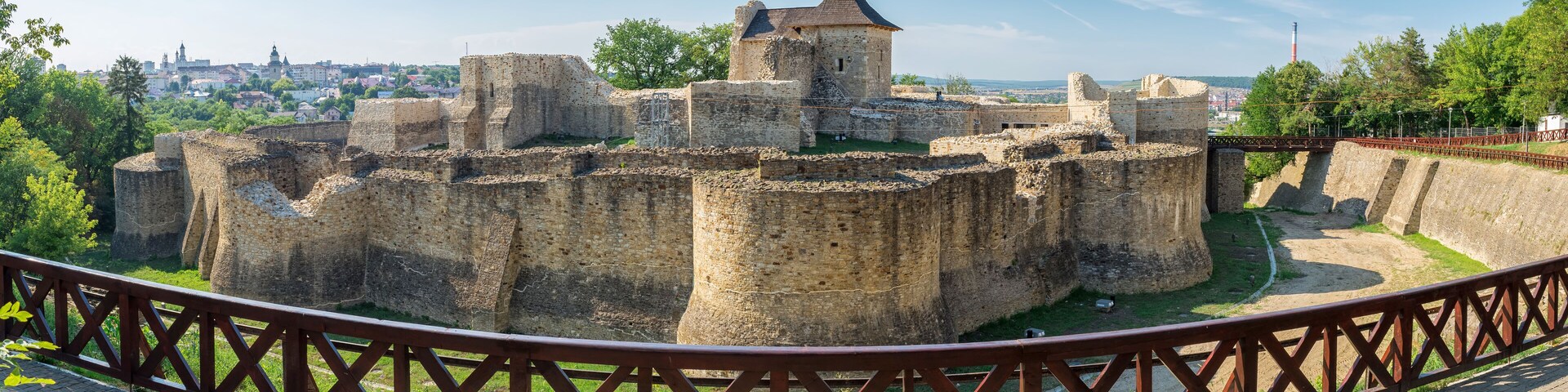A historic stone fortress with a central tower, surrounded by a moat and a wooden fence.