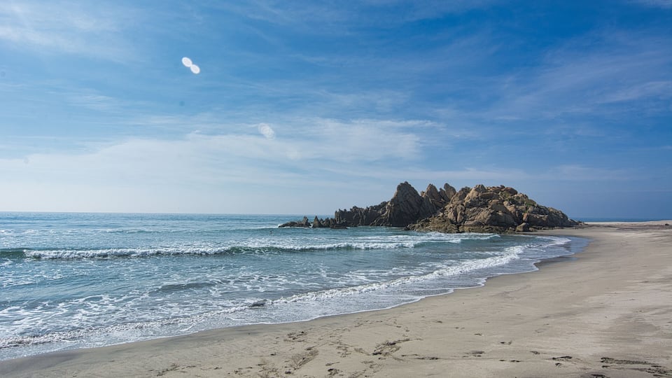 A sandy beach with waves, a rocky outcrop, and a clear blue sky.