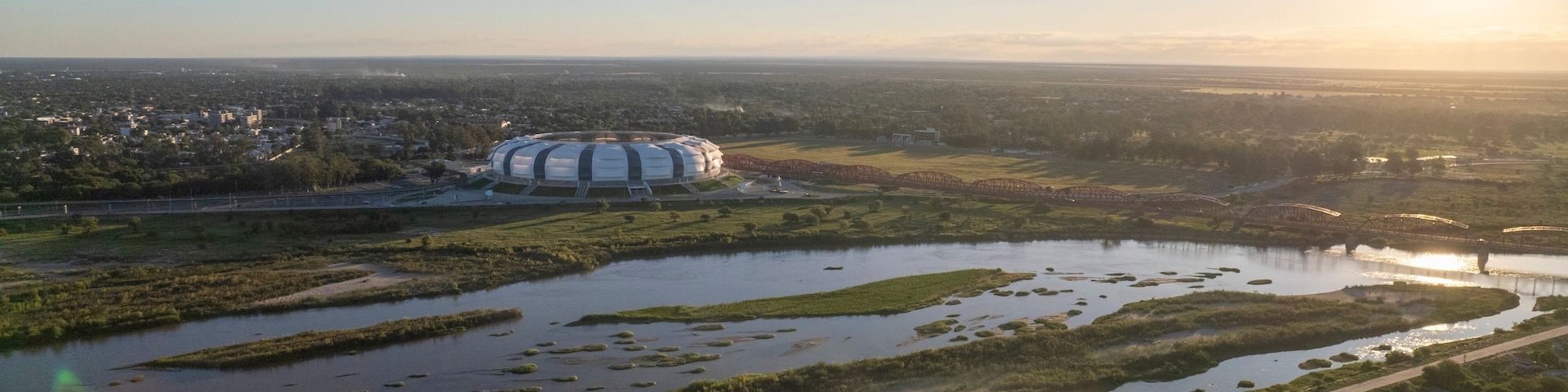 An aerial view of a city with a large stadium, a river, and surrounding greenery.
