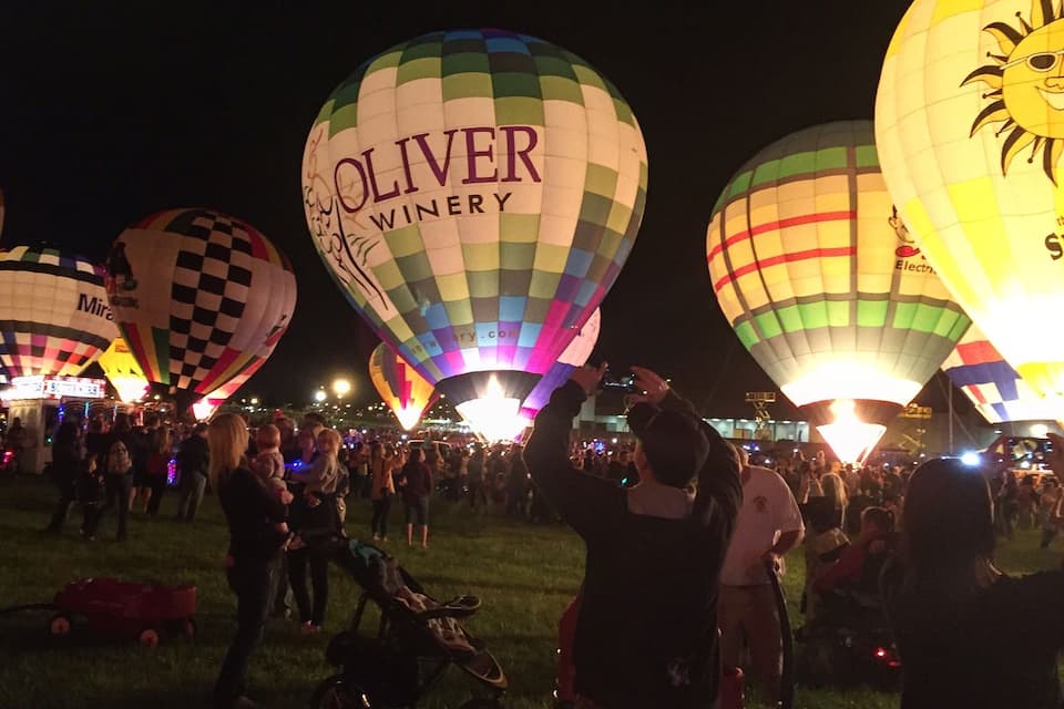 Um festival de balões de ar quente à noite, com balões coloridos, incluindo um com a inscrição 'OLIVER WINERY'.