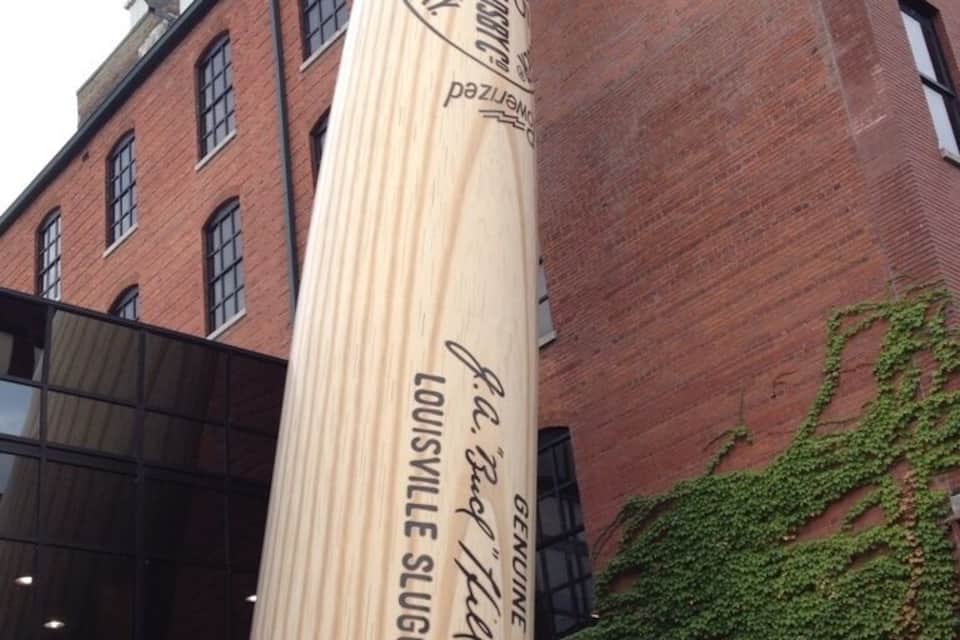 A Louisville Slugger baseball bat with the text "Louisville Slugger" and "Made in U.S.A." against a brick building with ivy.