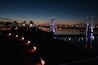 A nighttime cityscape with a lit bridge reflecting on the water.