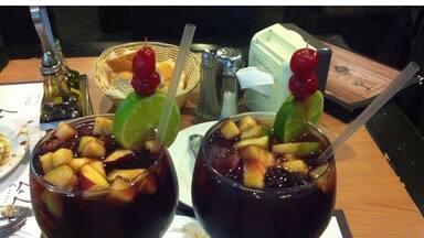 Two glasses of dark-colored beverage with fruit pieces and garnishes, on a table with a menu and a basket of bread.