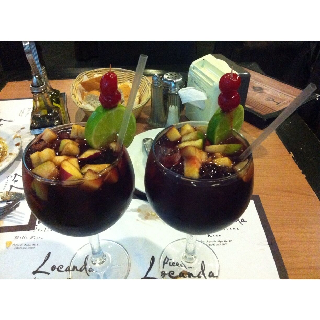 Two glasses of dark-colored beverage with fruit pieces and garnishes, on a table with a menu and a basket of bread.