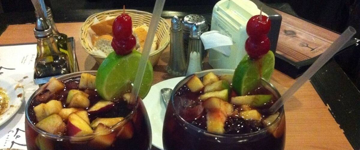 Two glasses of dark-colored beverage with fruit pieces and garnishes, on a table with a menu and a basket of bread.