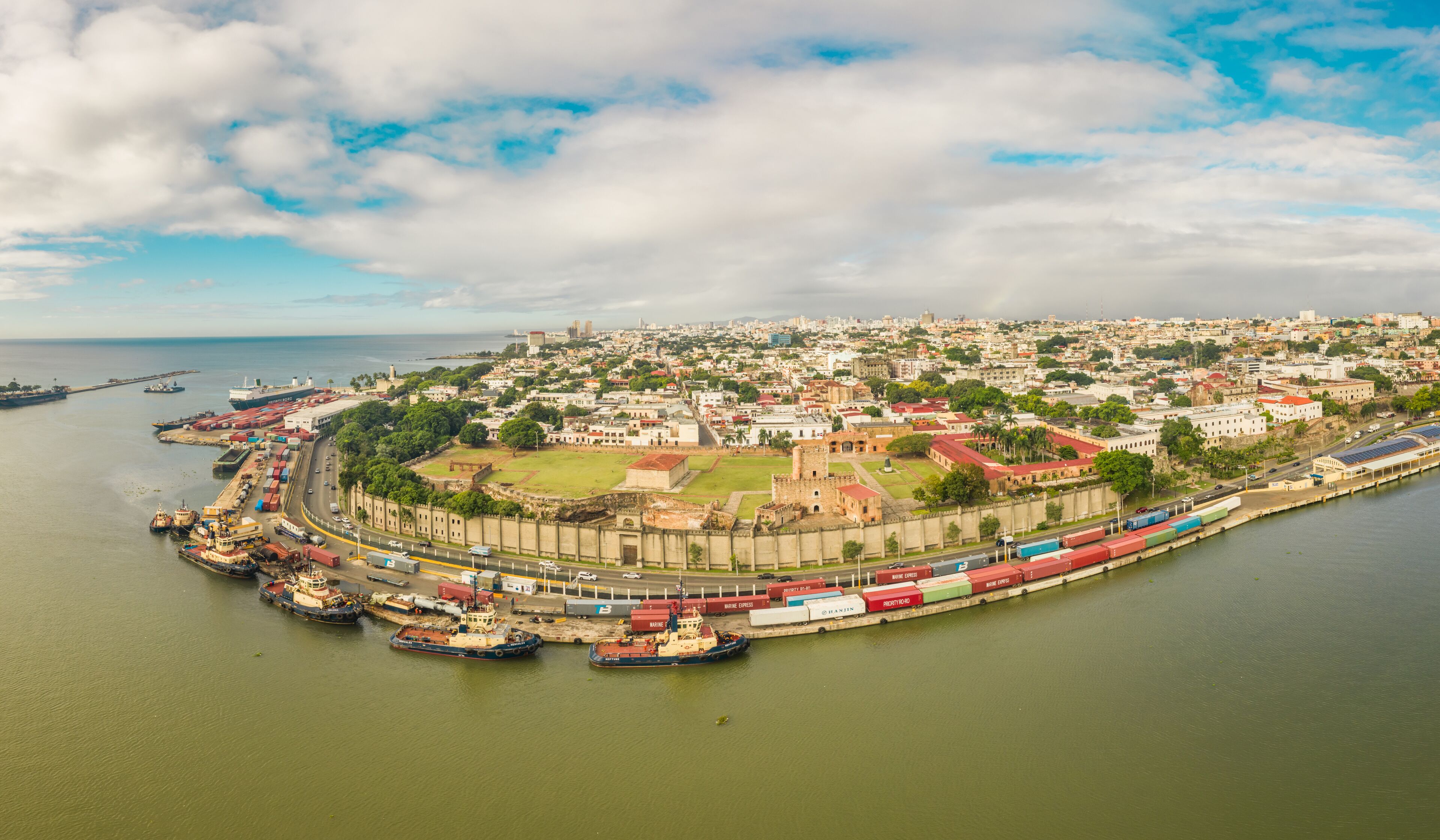 A coastal cityscape with a historic fort, a harbor with cargo ships, and a distant city skyline.