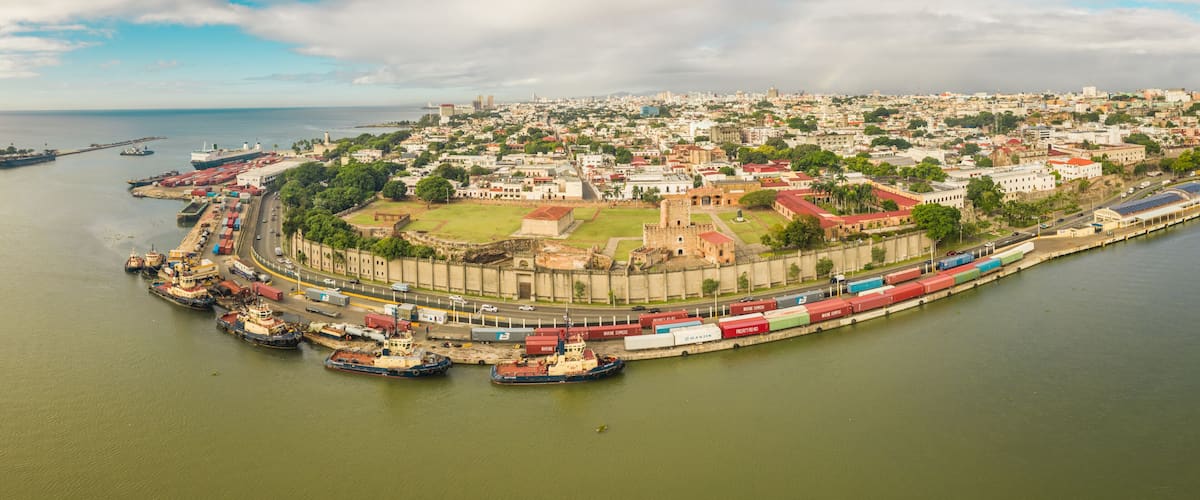 A coastal cityscape with a historic fort, a harbor with cargo ships, and a distant city skyline.