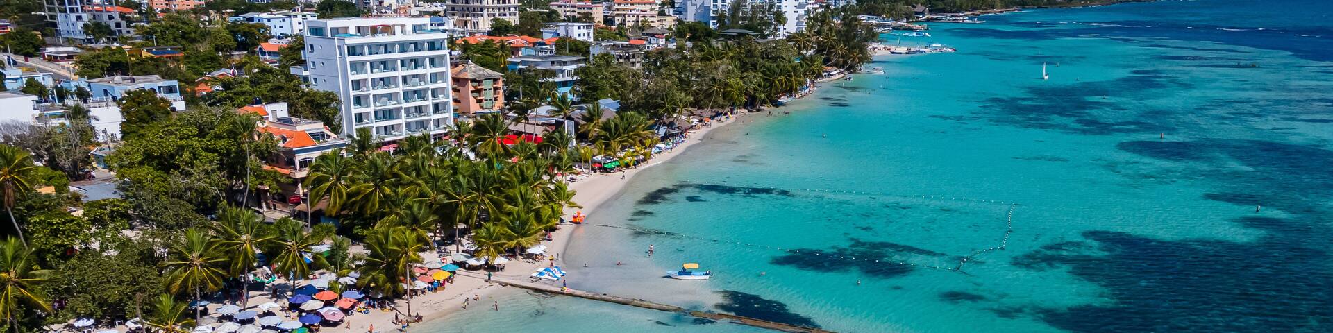 A coastal cityscape with a beach, clear turquoise waters, and palm trees.
