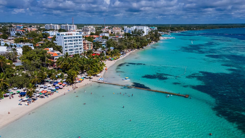 Un paisaje urbano costero con una playa, aguas turquesas cristalinas y palmeras.