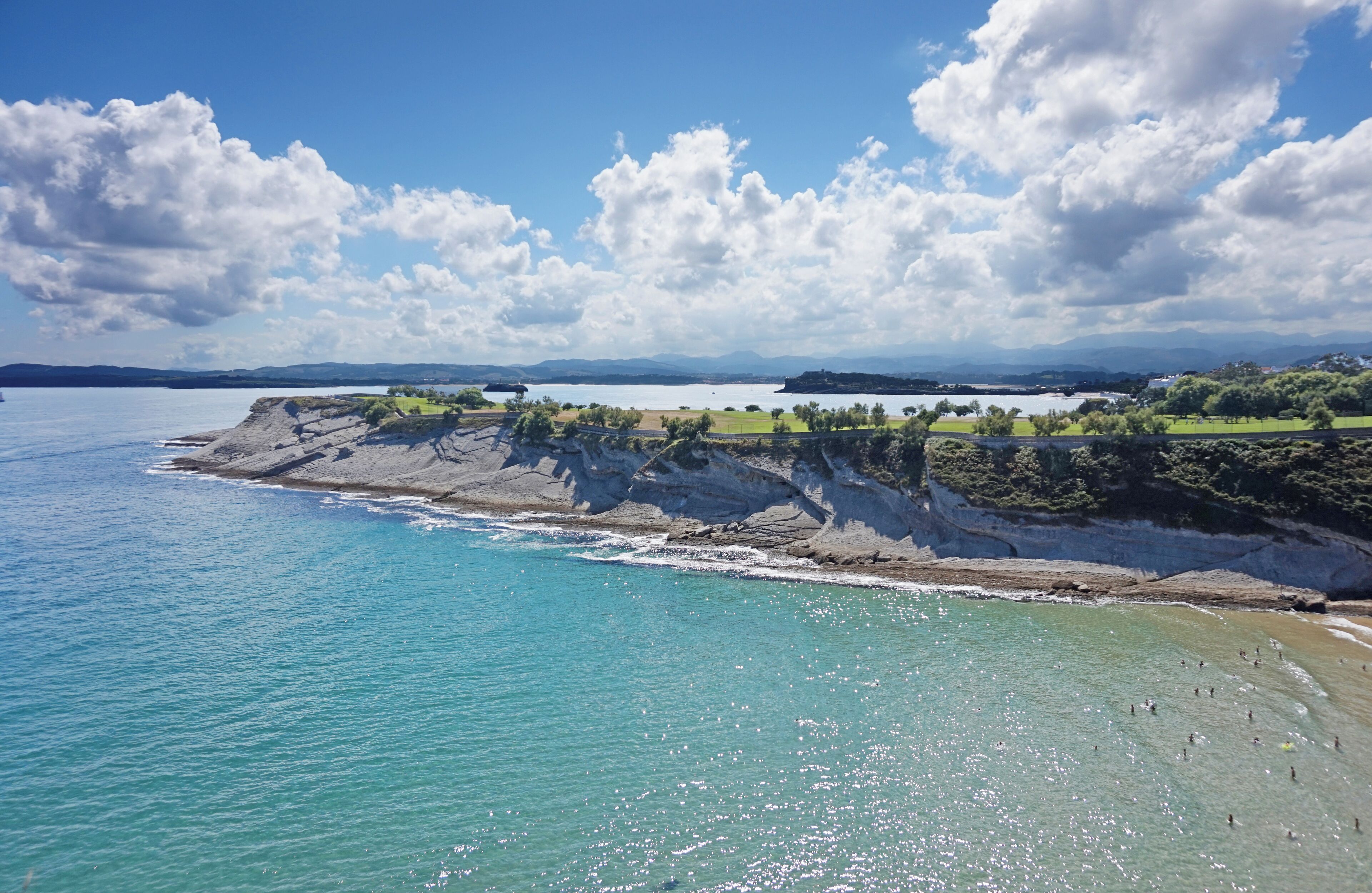 A coastal landscape with a clear blue sea, rocky cliffs, and a grassy area with trees.