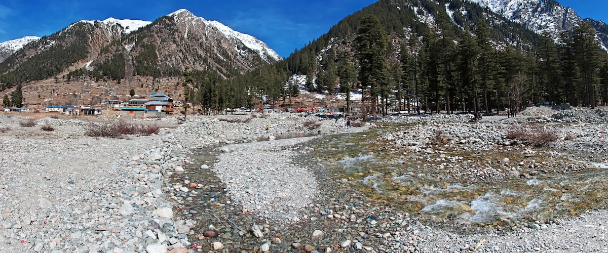 A mountainous landscape with a rocky stream, snow-capped peaks, and a small settlement with buildings.