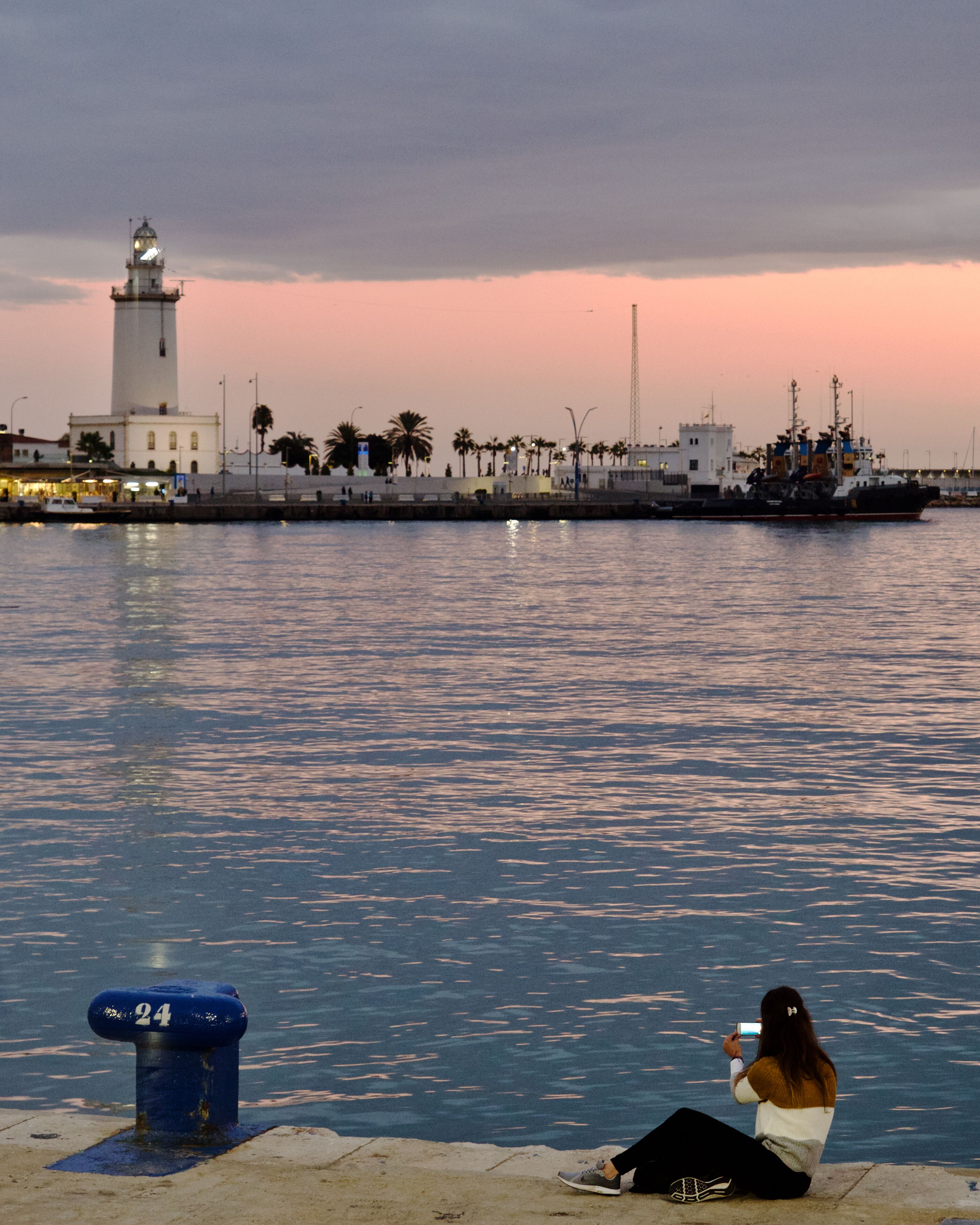 Girl photographing the lighthouse from the harbor promenade in Málaga, Spain.