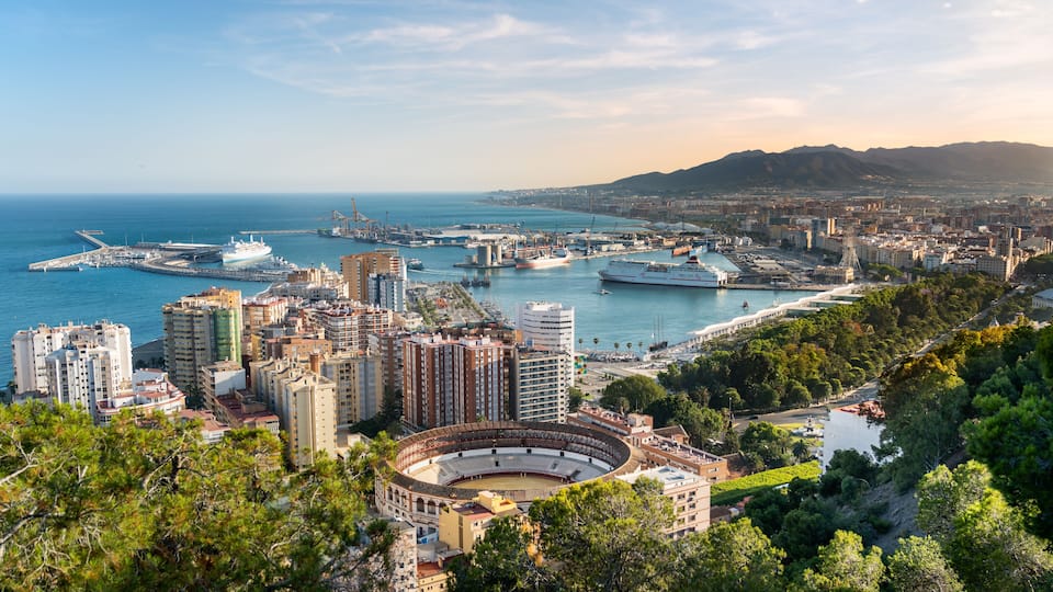 A coastal cityscape with a prominent amphitheater, a marina with ships, and a mountainous backdrop.