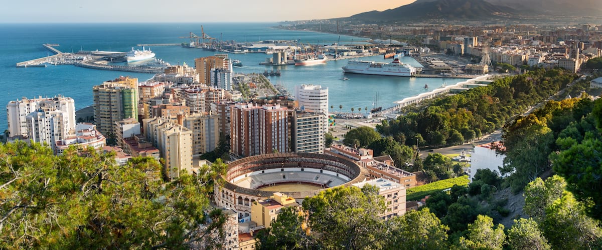 A coastal cityscape with a prominent amphitheater, a marina with ships, and a mountainous backdrop.