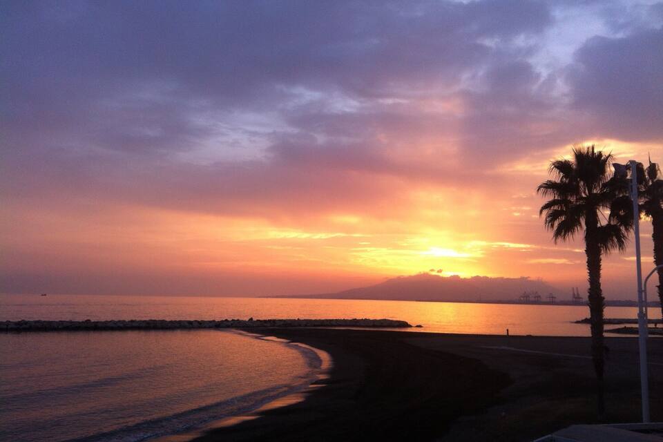 A sunset over a beach with palm trees and a rocky shoreline.