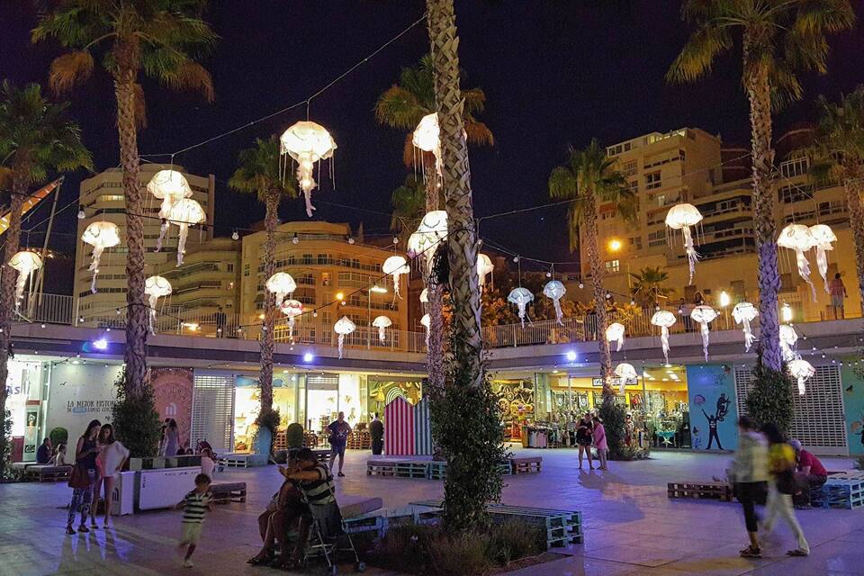 A plaza with palm trees, hanging lights, and people walking.