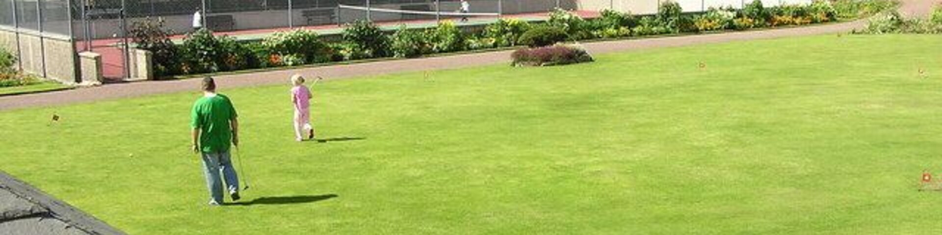 Un césped verde donde una persona y un niño juegan, rodeado de casas y una cerca.