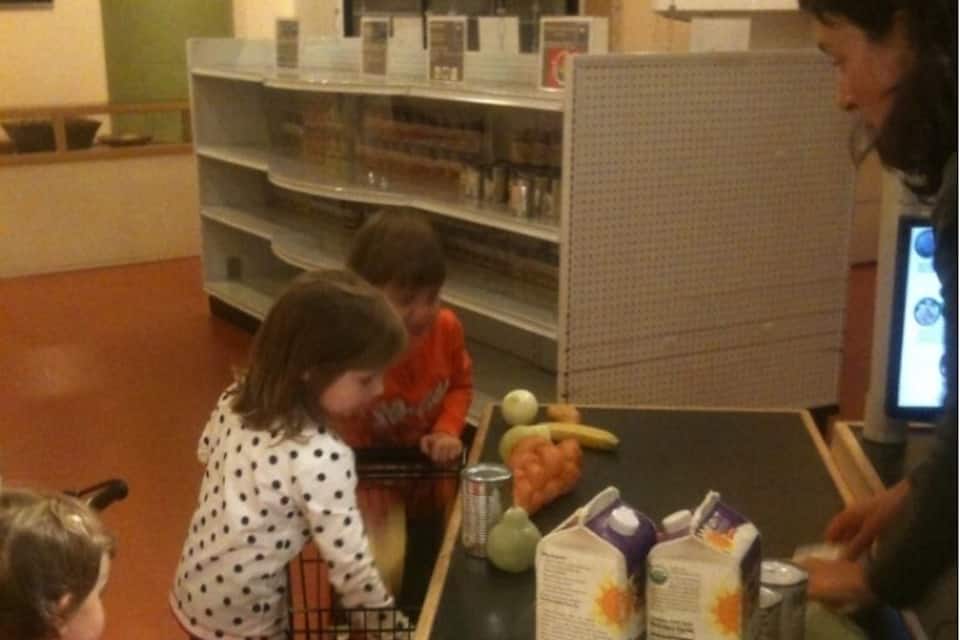 Un supermercado con niños y un adulto. Se ven estantes con productos y un refrigerador.