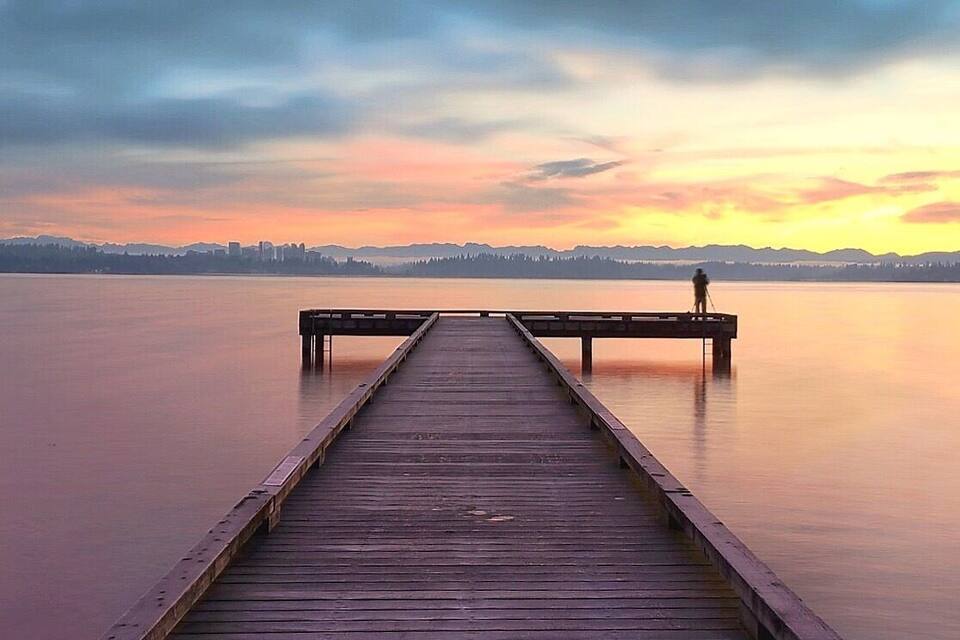 Een houten steiger die zich uitstrekt over kalm water, met een persoon aan het einde, uitkijkend op een stadszicht bij zonsondergang.