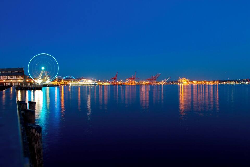 A waterfront at night with a Ferris wheel, cranes, and illuminated buildings reflecting on the water.