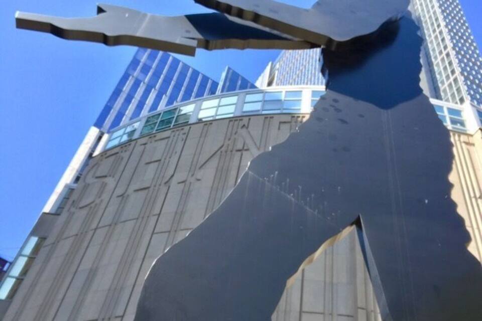 Una gran escultura metálica de una figura con brazos extendidos, frente a un fondo de edificios modernos y un cielo azul despejado.
