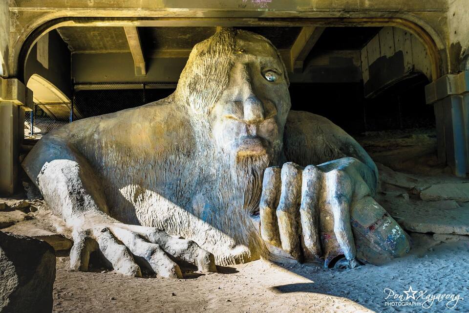 Een groot, verweerd beeldhouwwerk van een menselijke figuur met uitgestrekte armen en een baard, gelegen in een industriële omgeving met betonnen constructies en metalen balken op de achtergrond.