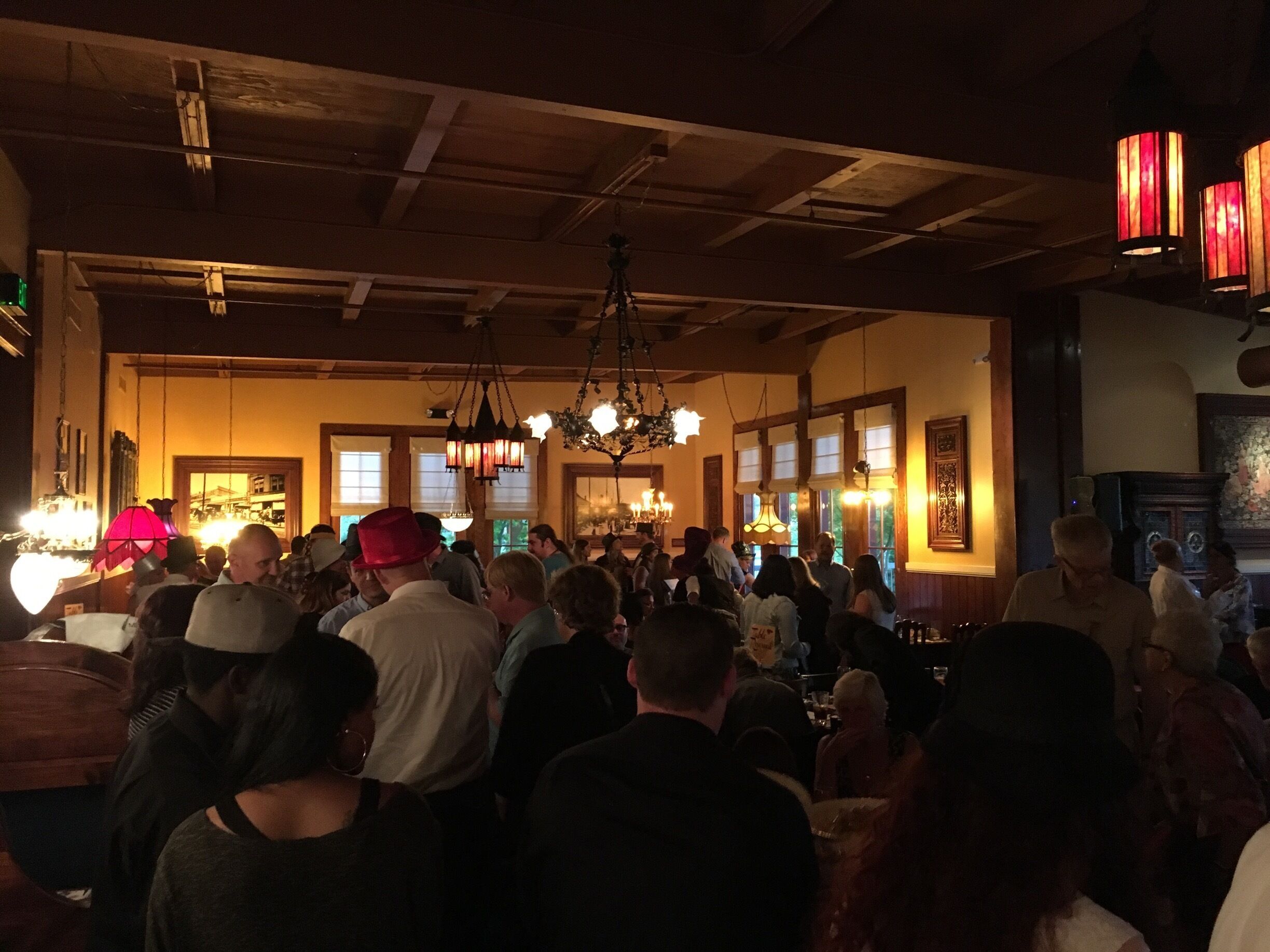 A crowded indoor venue with wooden beams, chandeliers, and large windows.