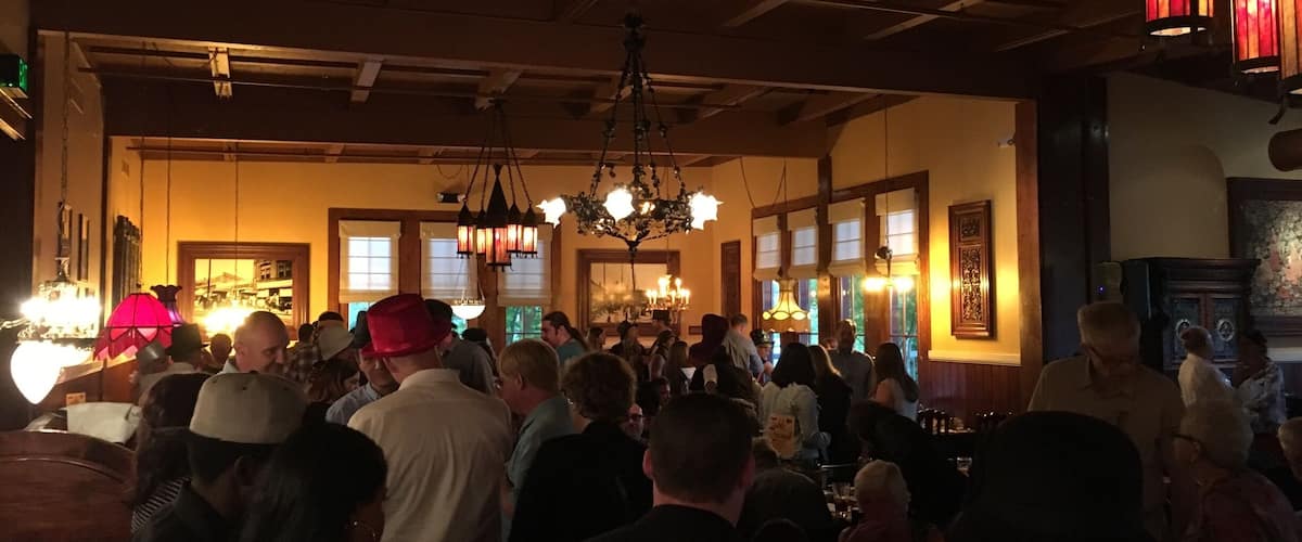 A crowded indoor venue with wooden beams, chandeliers, and large windows.
