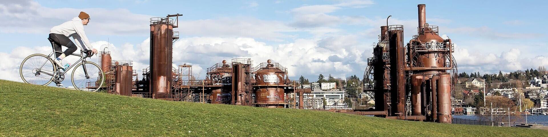 A person riding a bicycle on a grassy hill with industrial structures in the background.