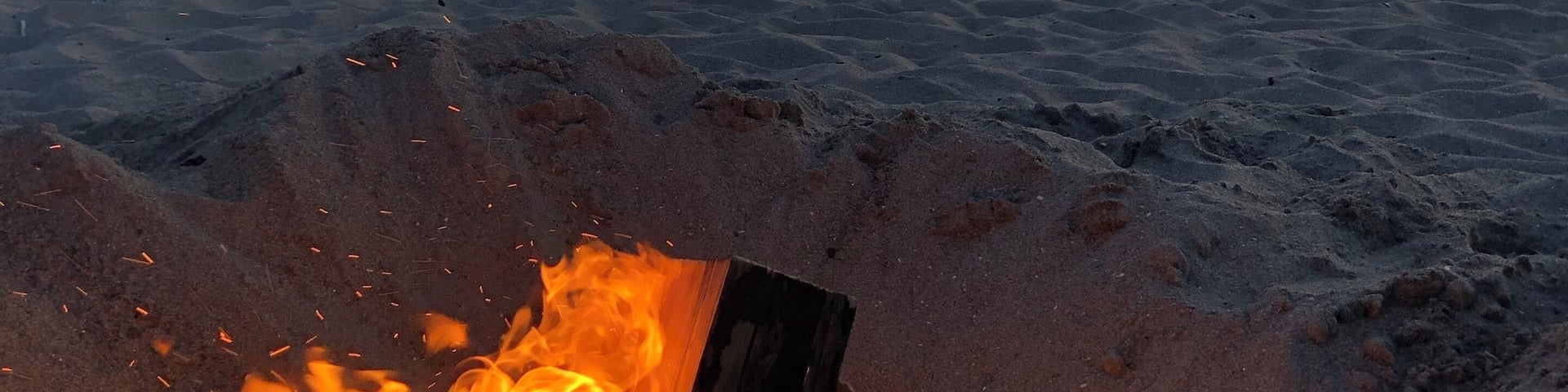 Una persona está de pie en la playa al atardecer, con una fogata al frente.