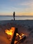 Een persoon staat op het strand bij zonsondergang, met een kampvuur op de voorgrond.