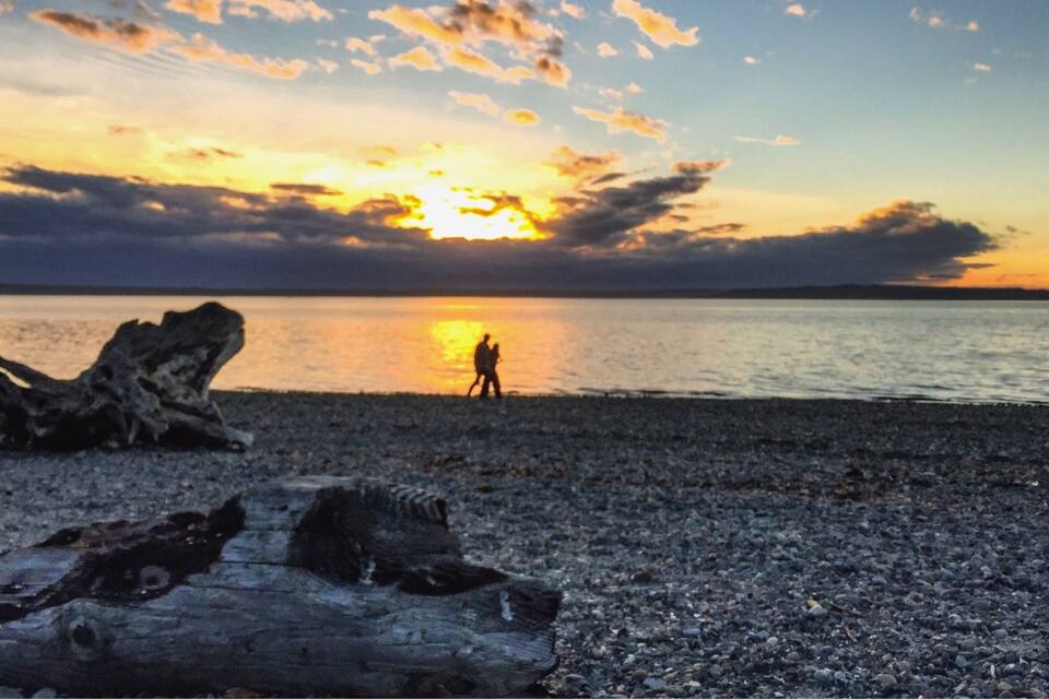Un atardecer sobre un mar tranquilo, con siluetas de personas y una gran formación rocosa en la orilla.