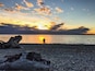 A sunset over a calm sea with silhouettes of people and a large rock formation on the shore.