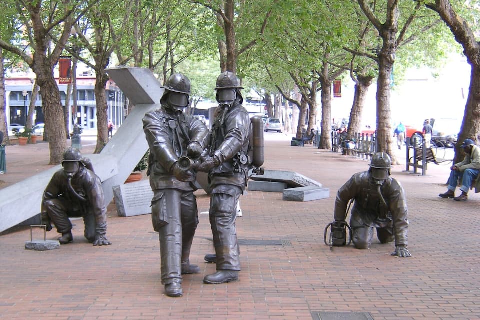 A bronze statue of three men in military gear, a large abstract sculpture, and a brick plaza with trees and benches.