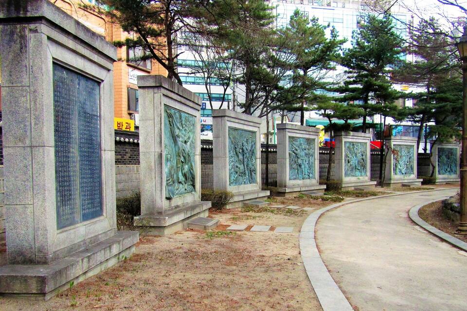 A row of stone monuments with inscriptions, surrounded by trees and a paved path.