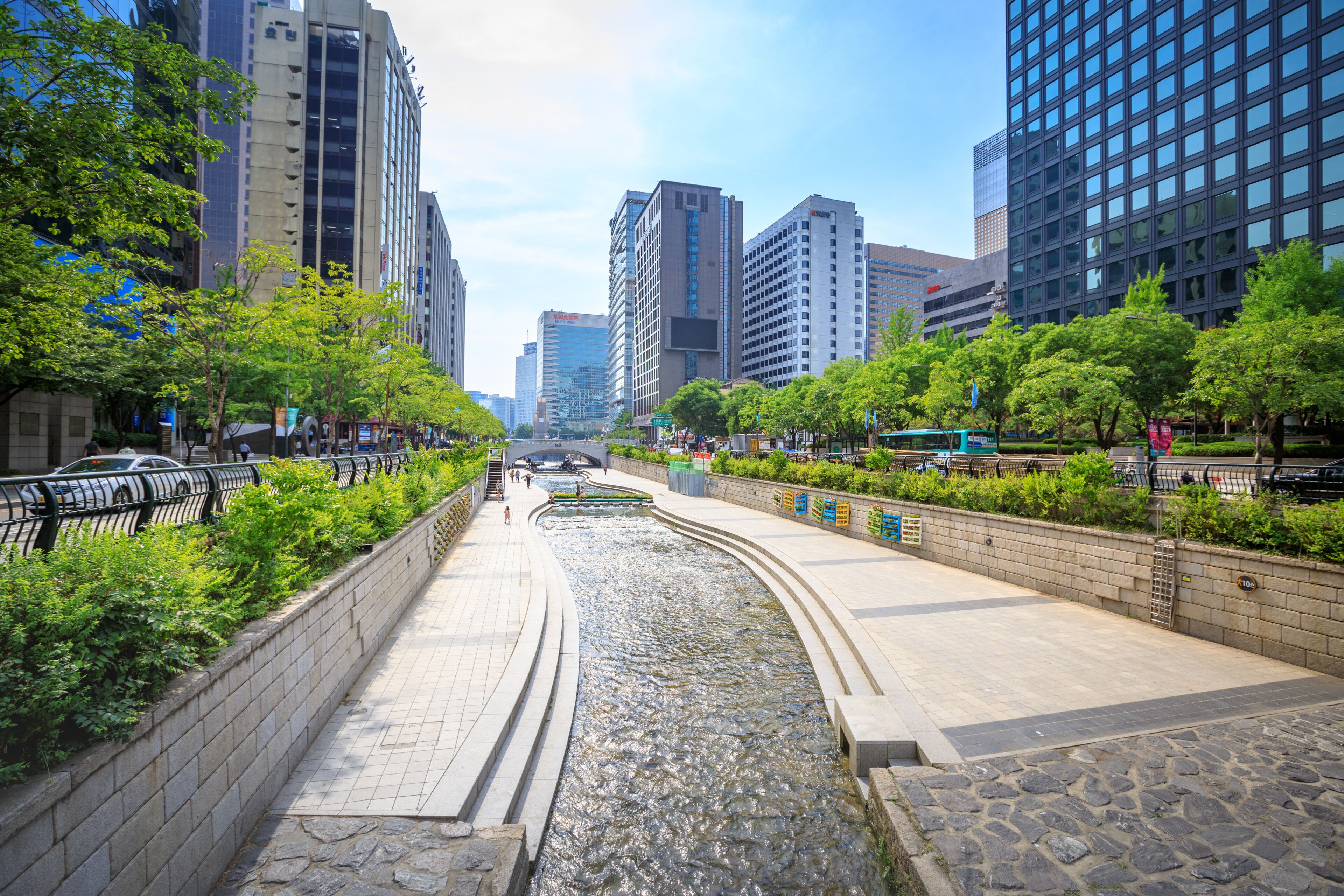 Un paisaje urbano con un río que atraviesa un paseo peatonal bordeado de árboles y edificios modernos.