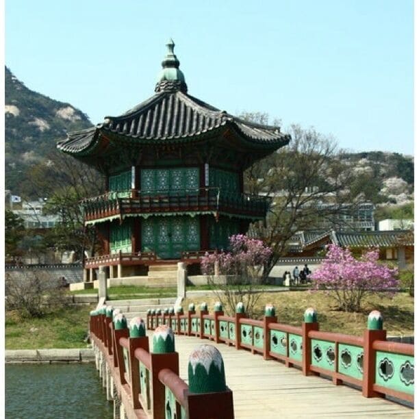 Un pabellón coreano tradicional con detalles ornamentados en verde y rojo, un puente de madera con barandillas decorativas y flores de cerezo al fondo.