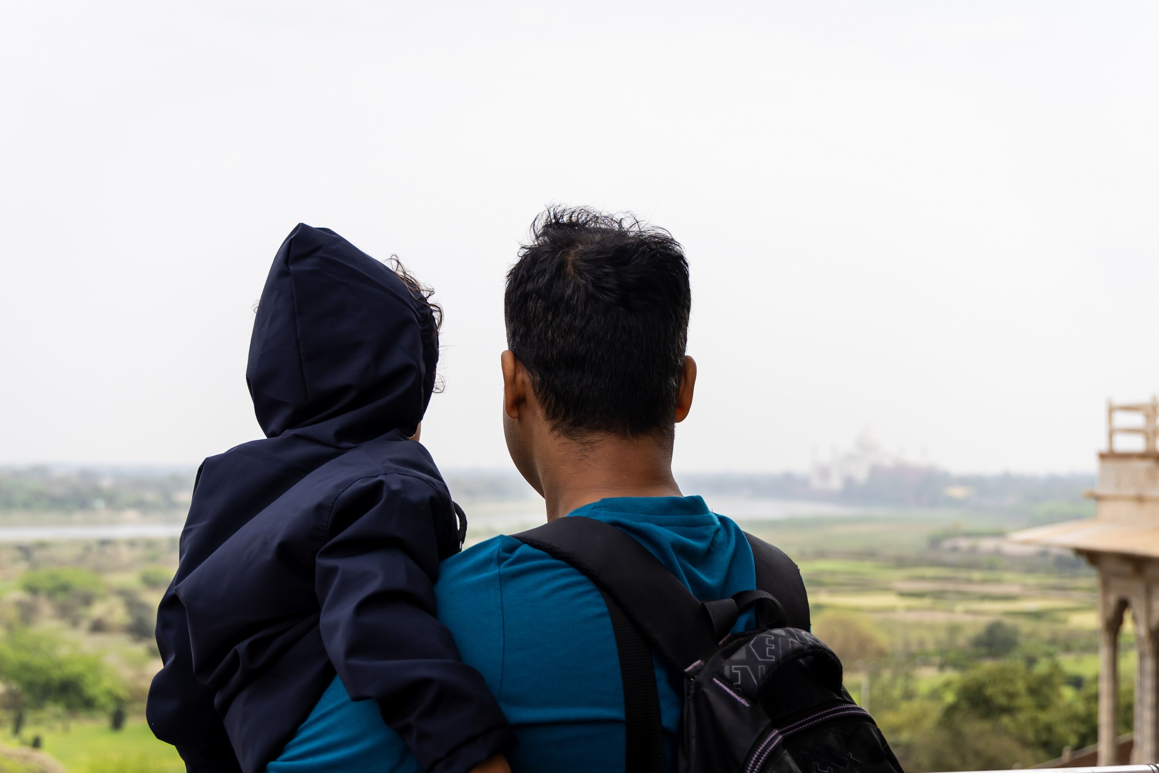 Dos personas, una cargando a la otra, contemplando un paisaje con campos y una estructura distante.