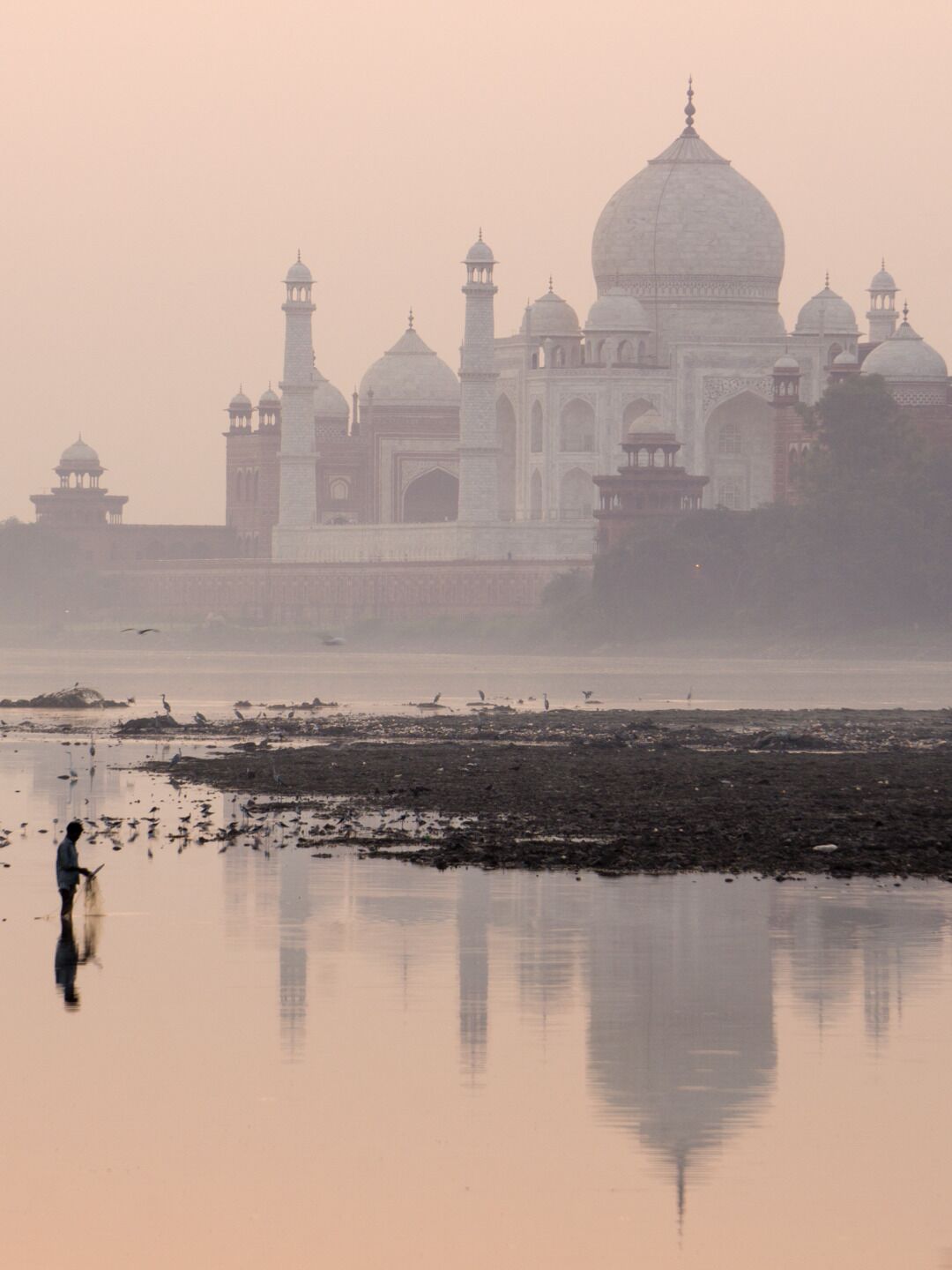 El Taj Mahal con su reflejo en el agua, una persona pescando y un cielo brumoso.