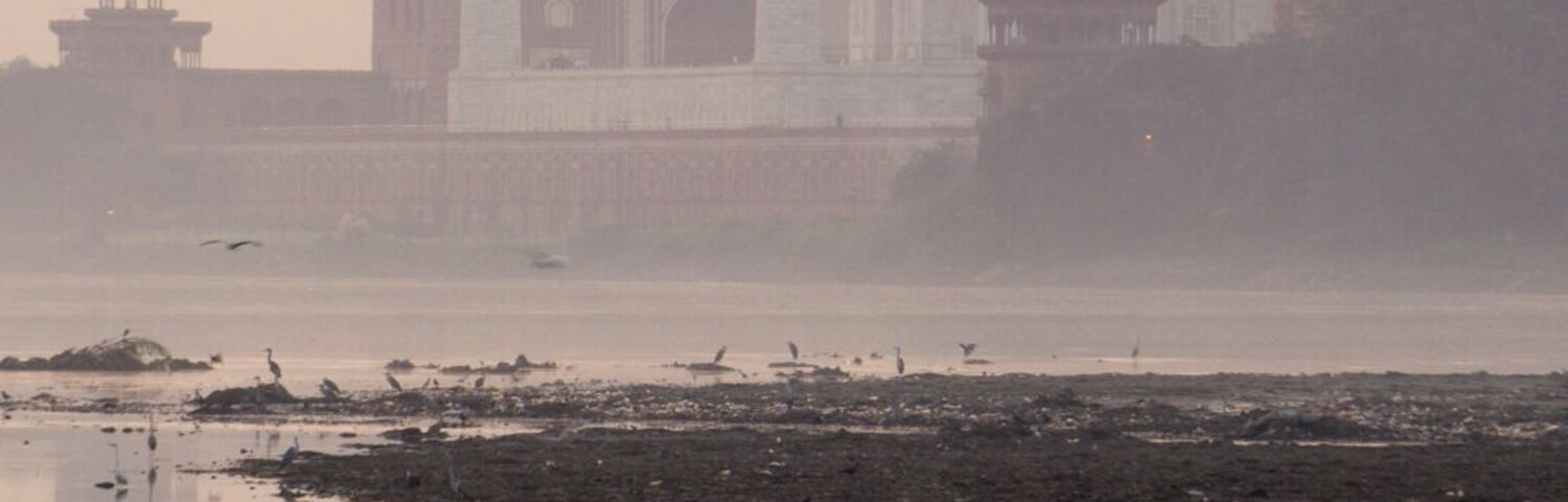 El Taj Mahal con su reflejo en el agua, una persona pescando y un cielo brumoso.