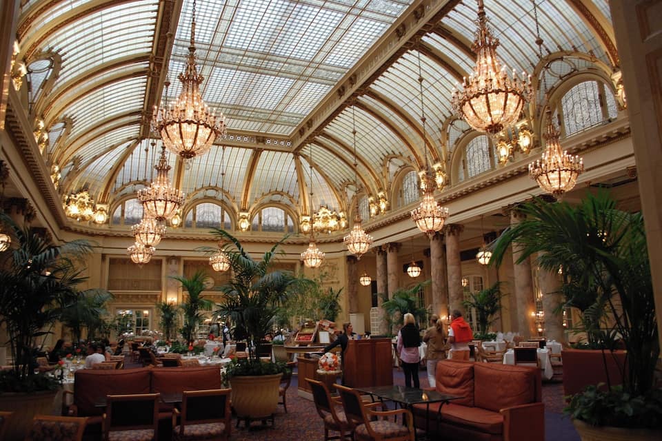 A grand, ornate hotel lobby with high ceilings, chandeliers, and lush greenery.