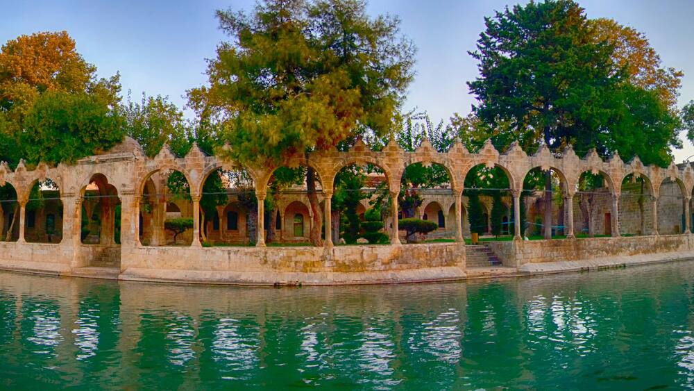 a peaceful panoramic view of balıklıgöl in şanlıurfa with historic arches, calm water reflecting the surrounding trees and mosque under a colorful sunset sky