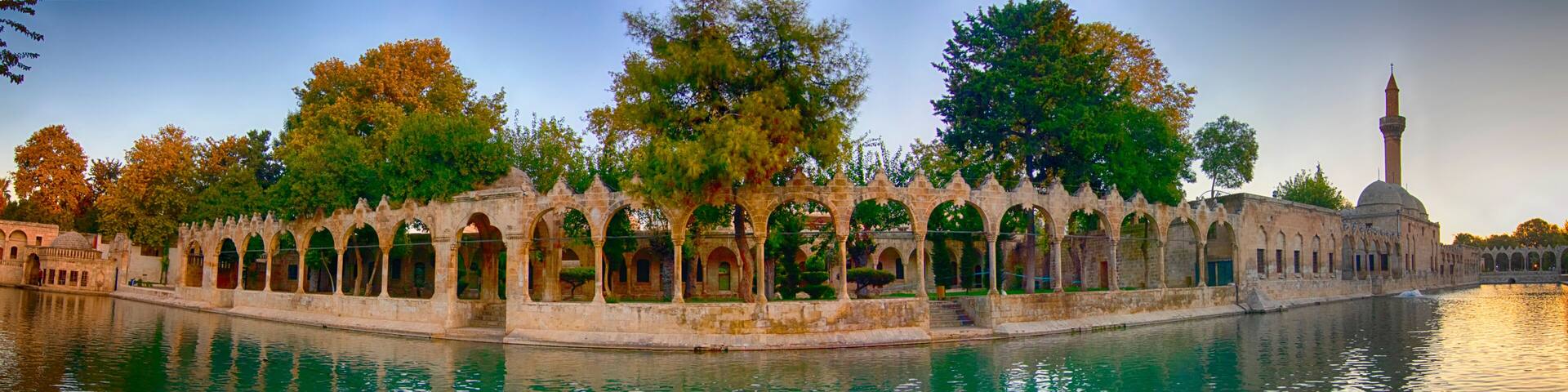 a peaceful panoramic view of balıklıgöl in şanlıurfa with historic arches, calm water reflecting the surrounding trees and mosque under a colorful sunset sky