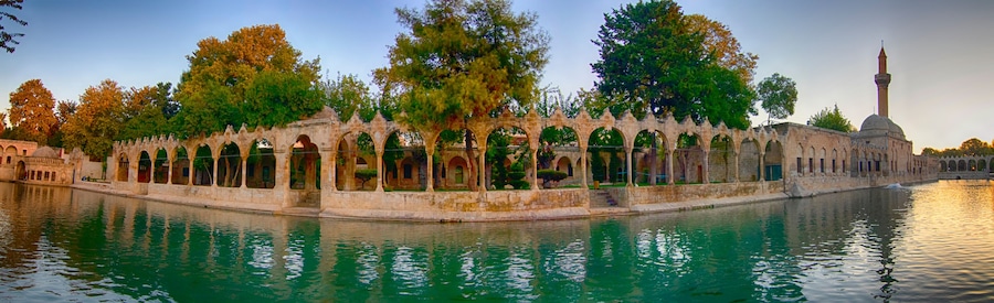 a peaceful panoramic view of balıklıgöl in şanlıurfa with historic arches, calm water reflecting the surrounding trees and mosque under a colorful sunset sky