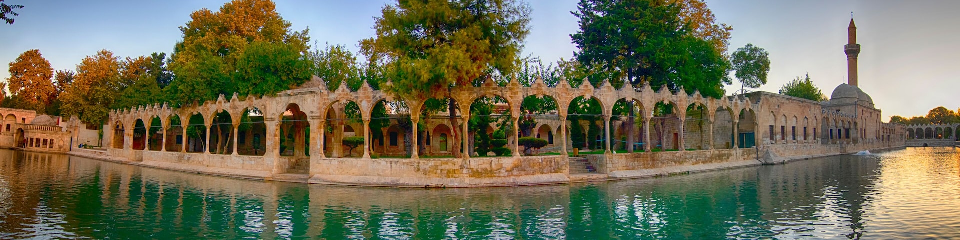 a peaceful panoramic view of balıklıgöl in şanlıurfa with historic arches, calm water reflecting the surrounding trees and mosque under a colorful sunset sky