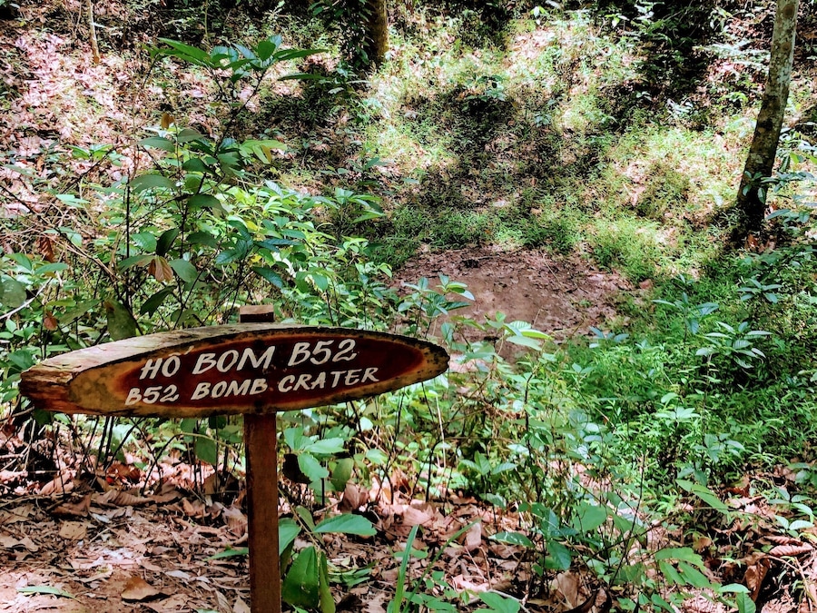 Ein Holzschild in bewaldeter Landschaft mit dem Text 'HO BOM B52 B52 BOMB CRATER'.