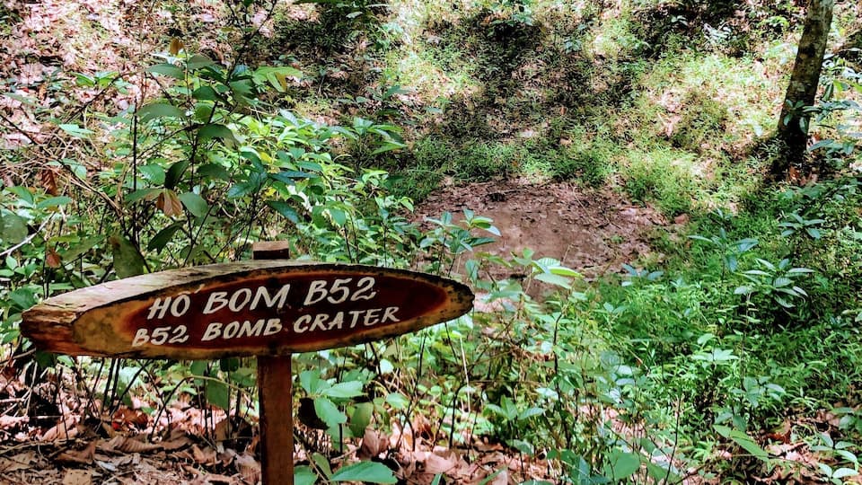 A wooden sign in a forested area with text reading "HO BOM B52 B52 BOMB CRATER".