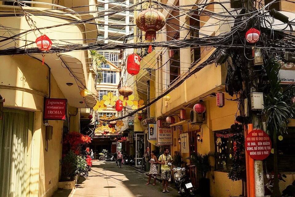 Uma rua estreita com edifícios amarelos, lanternas vermelhas e placas indicando um restaurante japonês e um hotel.